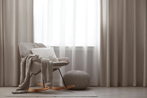 Modern cozy corner with a beige rocking chair, knitted pouf, and layered sheer and beige curtains filtering soft daylight.