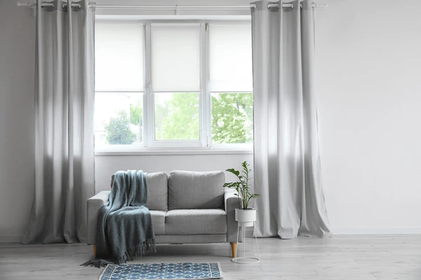 Modern Dubai living room with white walls, floor-length silver curtains, small grey sofa, indoor plant, and a patterned blue rug.

