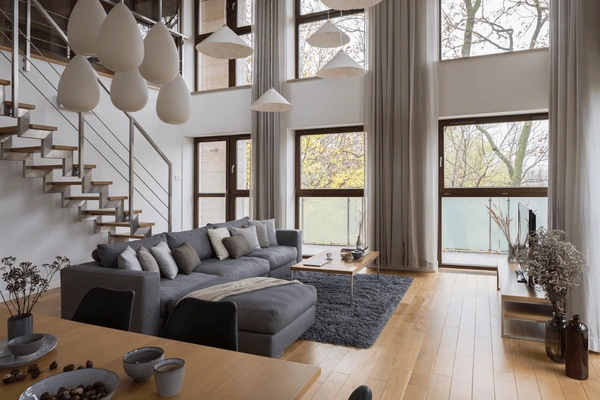 Stylish loft apartment with double-height windows, elegant falling curtains, modern grey sofa, and wooden flooring in Dubai 2025.