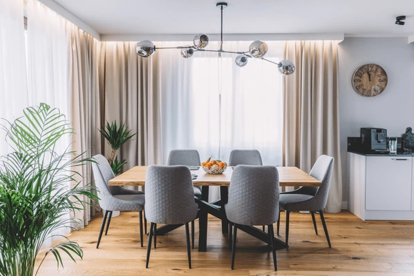 Apartment dining room with wooden table, gray chairs, and floor-to-ceiling beige long curtains enhancing a cozy modern interior.