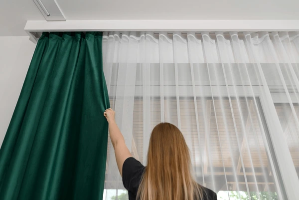 A woman adjusts elegant emerald green curtains layered with sheer white drapes, showcasing modern and affordable window treatment design for cozy Dubai homes in 2025.