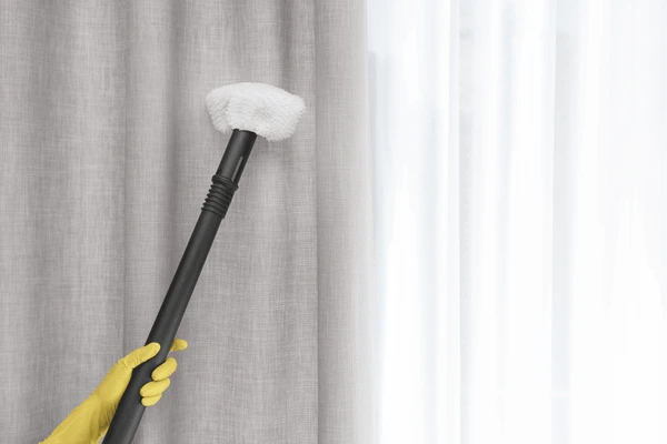 A person wearing yellow gloves cleaning light gray curtains with a steamer, representing easy-maintenance and machine-washable curtains for Dubai homes.