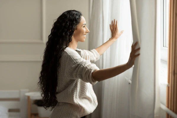 A smiling woman in a cozy modern Dubai home opening elegant white machine-washable curtains that softly filter natural sunlight into the room.