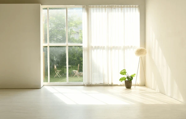 Bright minimalist living room in Dubai with white sheer measured curtains, soft sunlight, indoor plant, and large window view of garden chairs.