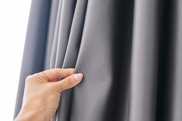 A close-up of a hand gently touching soft grey blackout curtain fabric, showing texture and quality. Best blackout curtains for Christmas in Dubai 2025 offering luxury and insulation.